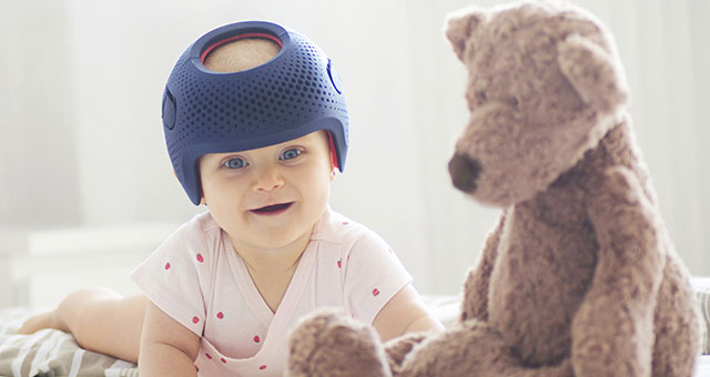 Baby trägt Talee-Helm und liegt auf dem Bauch, daneben sitzt ein Teddy